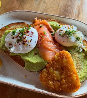 A close-up, overhead shot of a breakfast plate featuring poached eggs, smoked salmon, and avocado on toast, with an additional hash brown, served on a speckled ceramic plate on a wooden table.