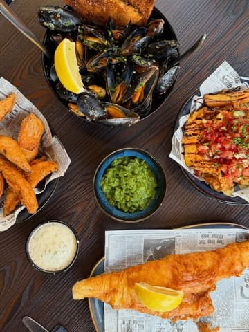 A high-quality, well-lit flat lay photograph of a seafood-focused meal featuring battered haddock, a bowl of mussels, dirty chips, sweet potato fries, and mushy peas, served on a dark wooden table with newspaper-style paper liners.