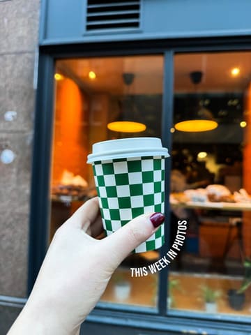 A standard, competent lifestyle shot featuring a branded coffee cup held in the foreground against a blurred, warm-lit cafe storefront. The composition is clean and the focus is sharp on the subject, effectively communicating a casual coffee-on-the-go moment.