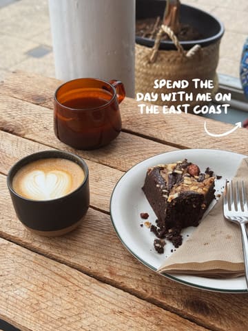 A high-angle, natural light shot of a coffee and a brownie on a rustic wooden table, likely taken at a cafe during a day trip. The image features a latte with heart art and a brownie with a bite taken out of it.