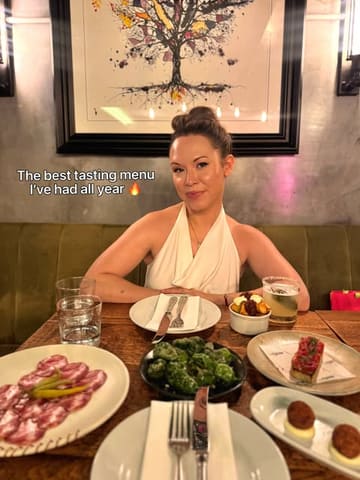 A photo of a woman sitting at a restaurant table filled with various small plates, including cured meats, padron peppers, and tartare. The image includes text overlay praising the tasting menu.