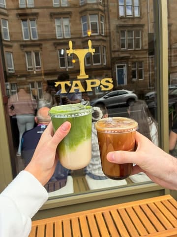 A casual, user-generated style photo showing two people holding takeaway drinks, specifically a matcha latte and an iced coffee, in front of a cafe storefront. The image captures a relaxed, on-the-go vibe typical of social media coffee recommendations.
