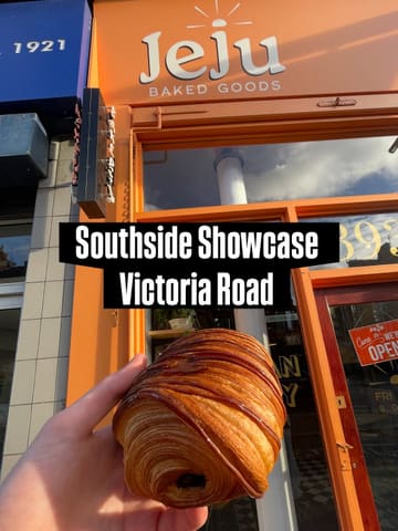 A handheld shot of a pain au chocolat held in front of the 'Jeju Baked Goods' storefront. The image serves as a visual hook for a local bakery showcase.