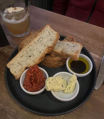A simple, overhead shot of a bread board starter featuring rustic bread, wild garlic butter, tomato tapenade, and balsamic oil, accompanied by a cocktail in the background.