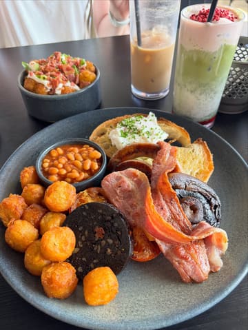 A high-quality, well-lit photograph of a hearty full English breakfast served alongside a side of tater tots and a vibrant matcha latte. The image captures the textures of the cooked breakfast items effectively, using natural light to highlight the freshness of the poached egg and the crispiness of the bacon.