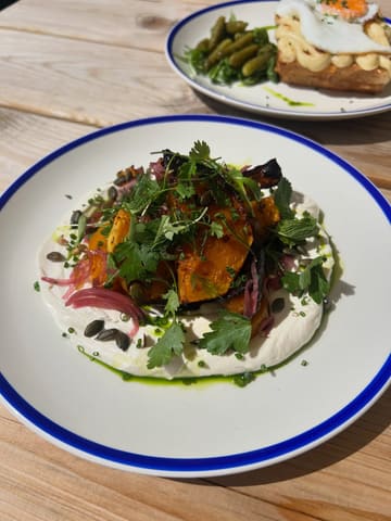 A bright, natural-light photograph of a roasted squash salad served on a bed of cream, topped with herbs and seeds, with a Croque Madame visible in the background.