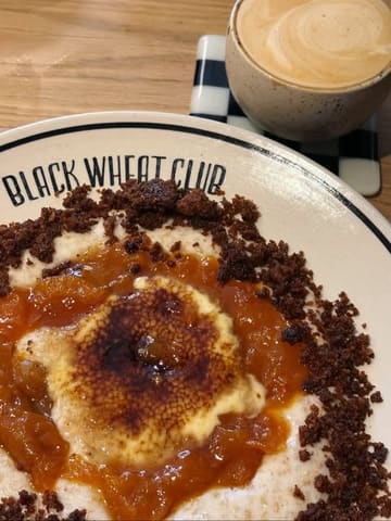 A high-quality, close-up shot of a creative 'Crème Brûlée Porridge' bowl served at Black Wheat Club in York. The dish features a distinct caramelized sugar top, fruit compote, and a crumble topping, presented on a branded plate alongside a flat white coffee. The lighting is soft and natural, highlighting the textures of the food.
