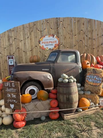 A bright, sunny, outdoor shot of a pumpkin festival display featuring a vintage truck, wooden barrels, and various pumpkins, set against a rustic wooden fence backdrop.