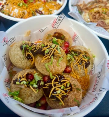A close-up, top-down shot of a bowl of Indian street food, specifically gol gappe, served in a branded paper-lined bowl. The image focuses on the textures and toppings of the dish.