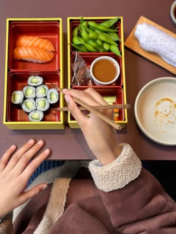 A clean, overhead, first-person perspective shot of a Japanese bento box meal featuring salmon nigiri, cucumber maki, and edamame, presented on a minimalist table setting.