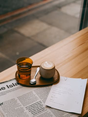 A high-quality, aesthetically pleasing shot of a flat white coffee served on a wooden table, accompanied by a menu and a newspaper. The composition uses a 45-degree angle to highlight the coffee cup, creating an inviting and cozy atmosphere typical of a specialty cafe.