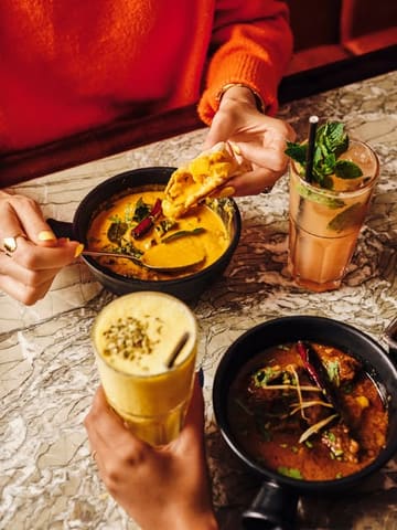 A high-quality, lifestyle-focused food photography shot featuring a spread of Indian dishes and drinks on a marble table. The image captures a social dining experience with hands interacting with the food, creating an inviting and warm atmosphere.