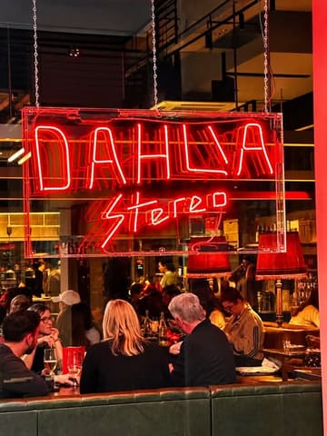 A moody, atmospheric shot of the interior of Dahlia Stereo in Manchester, focusing on the glowing neon sign above the dining area. The image captures the lively, social vibe of the venue with patrons in the foreground, emphasizing the nightlife and music-focused environment.
