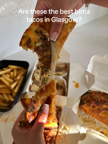A close-up, first-person perspective shot of a birria taco being pulled apart to showcase a cheese stretch, set against a backdrop of takeaway containers and fries on a marble surface.