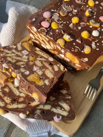 A high-quality, close-up shot of a homemade rocky road dessert topped with mini eggs. The lighting is natural and soft, highlighting the texture of the chocolate and biscuit chunks. The composition is effective, using a 45-degree angle to showcase the layers and the cross-section of the slice.
