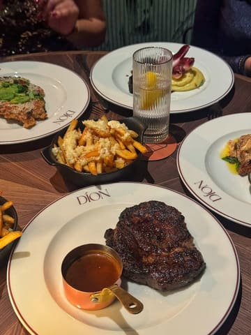 A top-down, slightly angled shot of a restaurant table spread featuring a rib-eye steak with a side of peppercorn sauce, truffle fries, and other dishes, capturing a group dining experience.