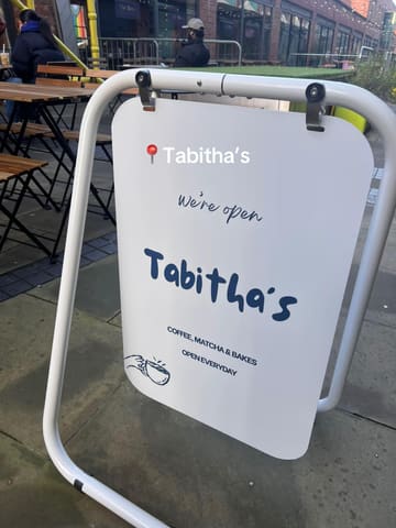 A functional, clear photograph of an A-board sign for 'Tabitha's' cafe, positioned outdoors. The image effectively communicates the venue's name and core offerings, though the composition is slightly compromised by the busy, cluttered background of the outdoor seating area.