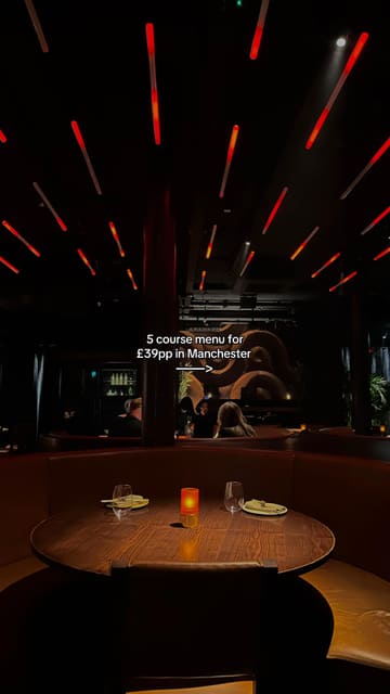 A moody, atmospheric shot of a restaurant interior, focusing on a table setting under ambient red lighting. The image effectively communicates the venue's vibe and the value proposition of a set menu.