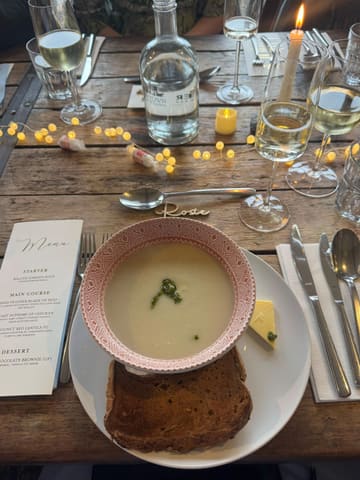 A high-quality, overhead flat-lay shot of a wedding table setting featuring a bowl of soup, a slice of bread, and a printed menu on a rustic wooden table. The scene is decorated with fairy lights and wine glasses, creating an elegant and inviting atmosphere.