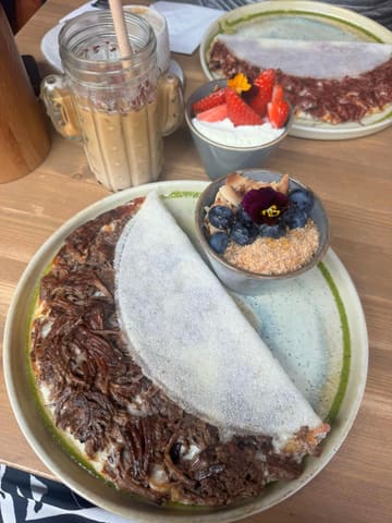 A bright, overhead shot of a Brazilian-style brunch featuring a beef brisket tapioca crepe, an acai bowl, and an iced latte. The lighting is natural and flattering, highlighting the textures of the food.