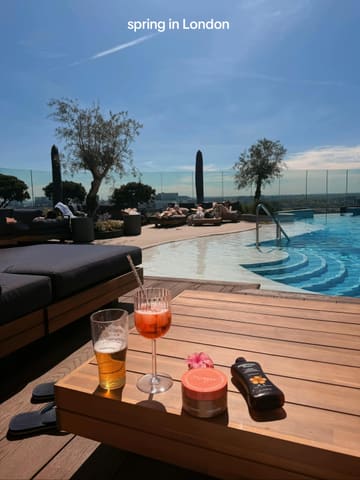 A lifestyle-focused shot taken on a rooftop terrace featuring a swimming pool, with a table in the foreground holding a beer, a cocktail, and tanning products. The image captures a sunny, relaxed atmosphere, though the inclusion of personal care items on the table detracts from the hospitality focus.
