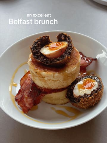 A high-angle, close-up shot of a savory brunch dish consisting of a stack of pancakes topped with black pudding and a soft-boiled egg, accompanied by crispy bacon and a drizzle of syrup, served on a white plate against a neutral grey background.