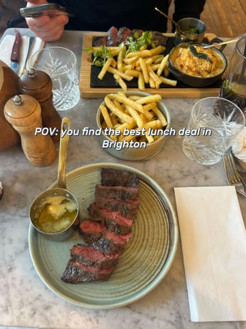 A POV-style overhead shot of a steak and fries lunch deal served on a marble table, featuring a side dish and drinks.