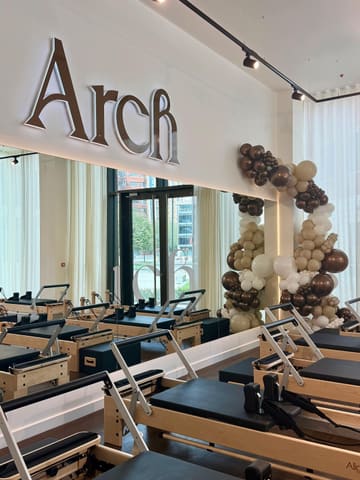 A bright, clean, and aesthetically pleasing shot of a Pilates studio featuring rows of reformer machines. The space is well-lit with natural light coming through the windows, and the decor is minimalist and modern, accented by a balloon arch.