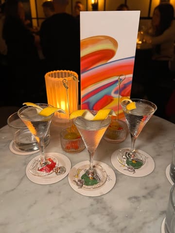 A close-up, eye-level shot of three 'Tiny Tini' cocktails served on a marble table in a dimly lit bar setting. The composition highlights the miniature nature of the drinks, accompanied by small bowls of garnishes and a candle, creating an intimate, atmospheric vibe.