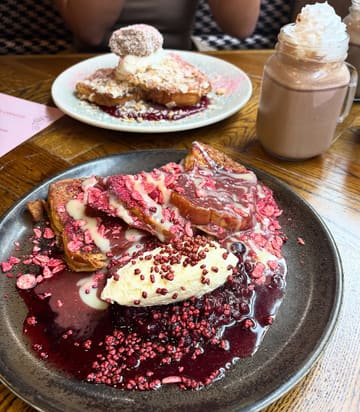 A high-quality, well-lit food photography shot featuring indulgent French toast dishes and a hot chocolate, likely taken at a brunch venue. The focus is sharp on the foreground plate, highlighting the textures of the toppings, while the background elements provide context without distracting from the main subject.