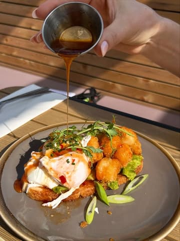 A high-quality, well-lit action shot of a brunch dish being drizzled with sauce. The composition is effective, capturing the texture of the food and the motion of the pour, making it highly appealing.