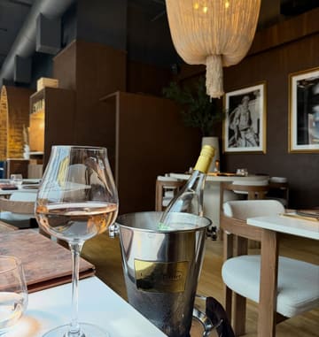 A well-composed, professional-looking shot of a restaurant table setting featuring a glass of wine and a bottle of Laurent-Perrier champagne in an ice bucket. The image captures an elegant, inviting atmosphere with a shallow depth of field that keeps the focus on the drinks while providing a soft, contextual view of the restaurant interior.