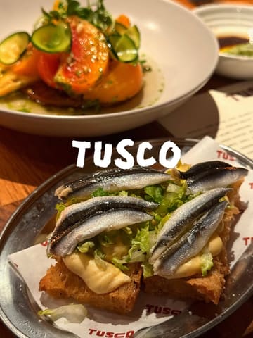 A close-up, appetizing shot of a parmesan Caesar crostini topped with white anchovies, with an heirloom tomato dish visible in the background, captured at a restaurant called Tusco.