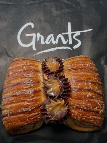 A high-quality, top-down close-up shot of a laminated pastry, likely a danish or croissant-style treat, featuring piped chocolate and caramel ganache. The pastry is placed on a branded paper bag from 'Grants', creating a clean, professional product shot that highlights the intricate layers and textures of the bake.