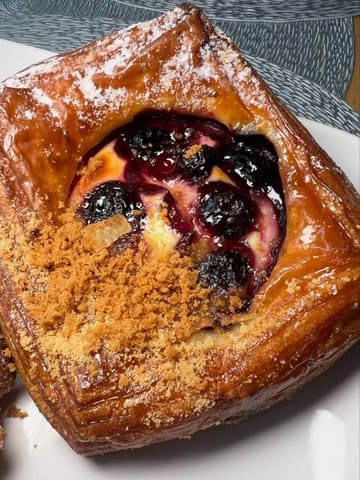 A high-quality, close-up photograph of a blueberry cheesecake danish pastry, highlighting the flaky texture and fruit filling.
