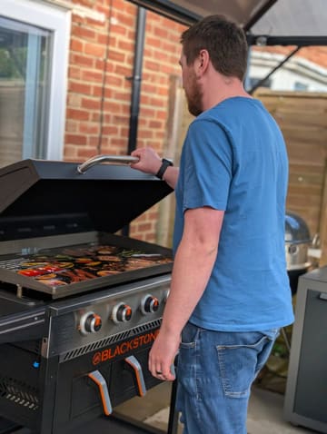A photo of a man standing in a garden, cooking on a Blackstone griddle. The shot is taken from behind the subject, focusing on the griddle surface which contains indistinct, out-of-focus food items.