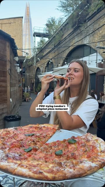 A POV-style shot of a person eating a large pizza at an outdoor venue in London, with The Shard visible in the background.