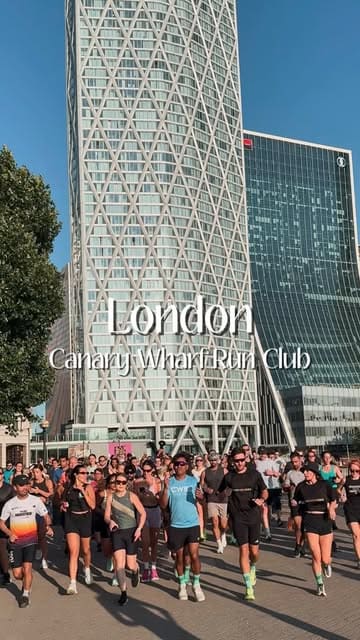 A bright, high-quality shot of a large group of people running together in Canary Wharf, London, promoting a local run club. The image captures a sense of community and active lifestyle against the backdrop of the city skyline.