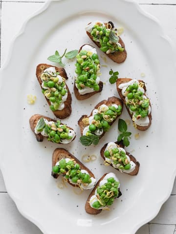 A high-quality, overhead food photography shot featuring several crostini topped with ricotta, smashed peas, pine nuts, and lemon zest on a white platter. The lighting is bright and even, highlighting the vibrant green of the peas and the creamy texture of the ricotta, creating a fresh and inviting aesthetic suitable for a Mediterranean-inspired menu.