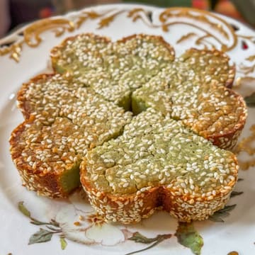 A high-quality, overhead close-up shot of a homemade Bingka Pandan, a traditional Malaysian kuih. The dish is presented on a decorative plate, clearly showcasing its vibrant green, custardy interior, caramelized crust, and a generous topping of toasted sesame seeds.