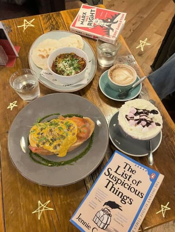 A cozy, overhead lifestyle shot of a cafe table featuring a variety of food and drinks alongside books, capturing a relaxed reading date atmosphere.