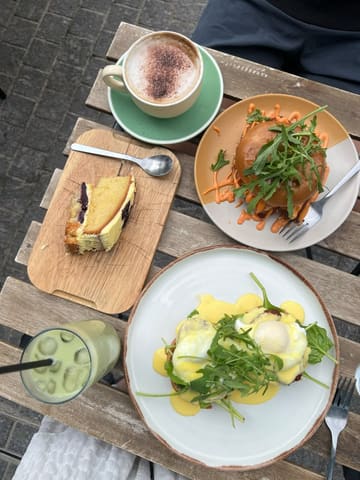 A top-down flat lay photograph of a brunch spread on an outdoor wooden table, featuring eggs benedict, a bagel, a slice of cake, a coffee, and an iced matcha latte.