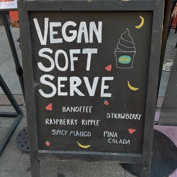 A clear, eye-level photograph of a chalkboard sign placed on a pavement outside a cafe, advertising vegan soft serve ice cream with a list of available flavors.