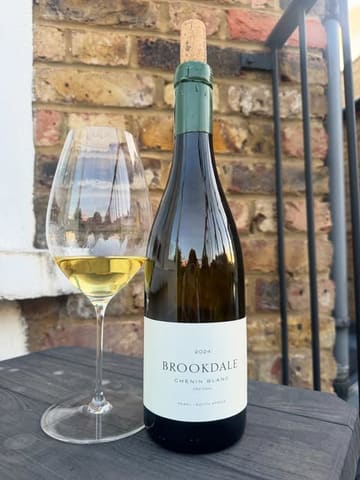 A simple, amateur-style photograph of a bottle of Brookdale Chenin Blanc and a glass of white wine, placed on a dark surface against a brick wall.