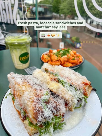 A top-down, slightly angled shot of a lunch spread featuring a chicken parm focaccia sandwich, a bowl of rigatoni, and a matcha latte, set on a green table in a food hall environment.