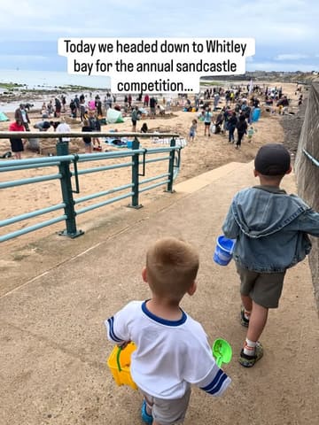 A candid, family-oriented shot capturing children walking down to a beach for a sandcastle competition. The image effectively conveys the atmosphere of a community event, though the background is visually busy.