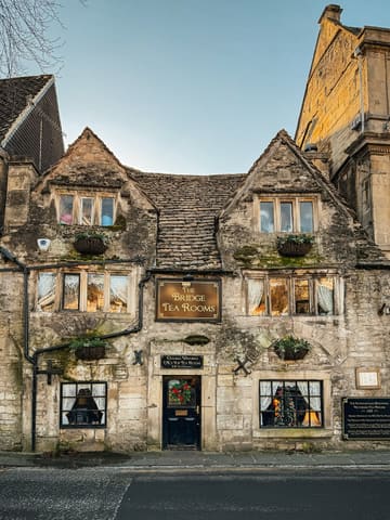 A high-quality, eye-level architectural shot of The Bridge Tea Rooms in Bradford on Avon. The image captures the historic, stone-built facade of the 1502 building during what appears to be golden hour, highlighting its traditional and nostalgic aesthetic.