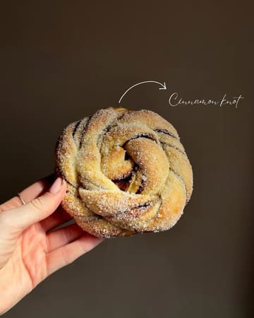 A high-quality, studio-style close-up shot of a cinnamon knot held in a hand against a dark, minimalist background. The lighting is soft and directional, perfectly highlighting the texture and sugar coating of the pastry, making it look fresh and appealing.