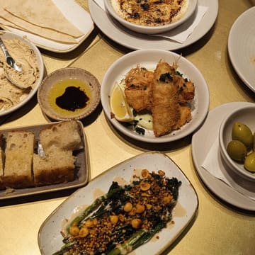 A flat-lay photograph of a tapas spread on a gold-colored table, featuring various dishes including olives, focaccia, hummus, charred broccoli, and crispy sea bass.