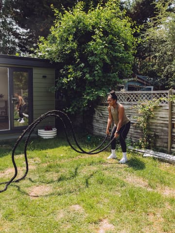 A woman performing battle rope exercises in her garden, marking the 14th anniversary of her fitness business.